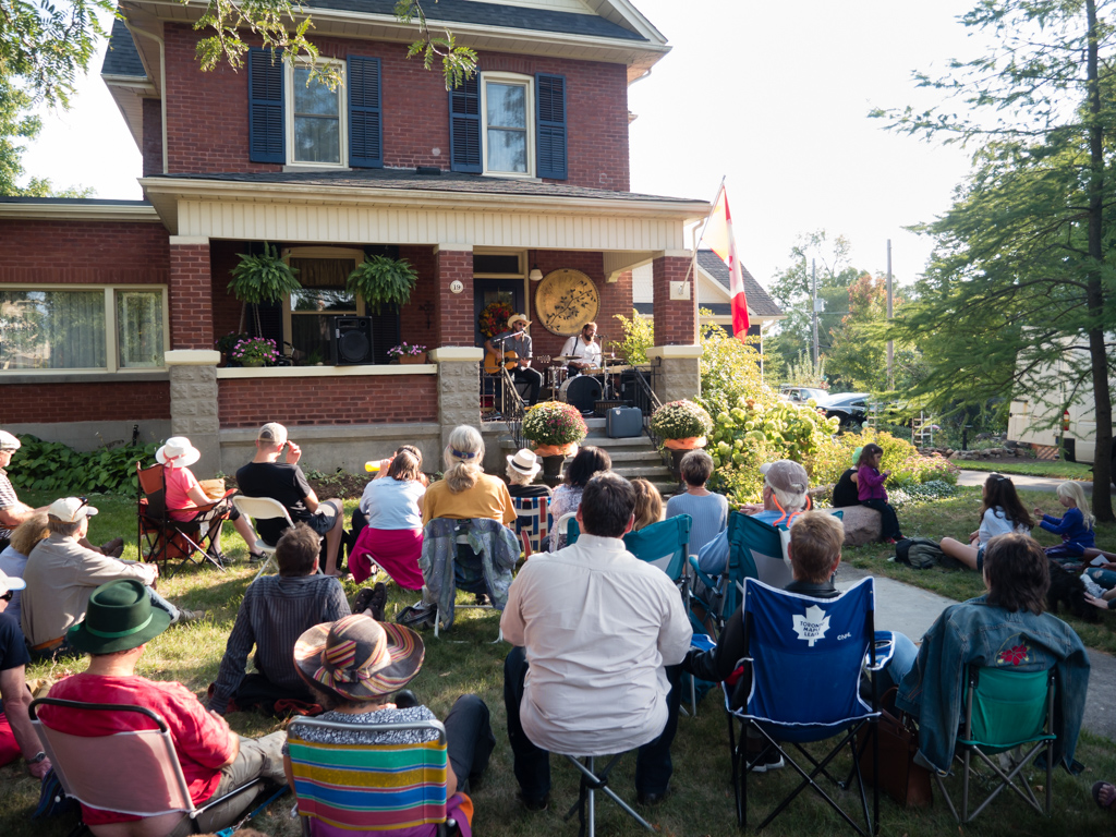 Musicians perform on a porch, with an audience sprawled out over the lawn